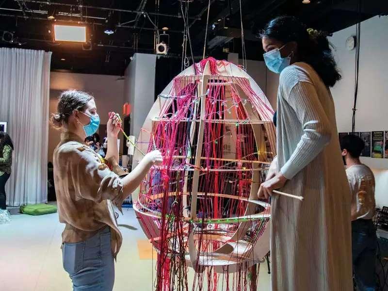 2 students work on a large, egg-shaped object weaving colored yarn